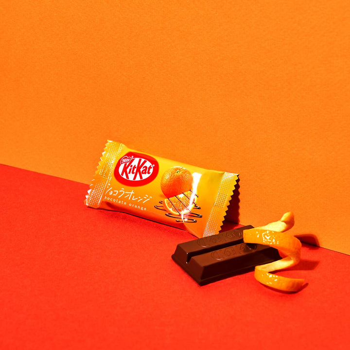 A Nestle Japan Chocolate & Ehime Iyokan Orange Kit Kat on a red and orange backdrop with an orange peel next to the open wrapper displaying a dark chocolate bar.