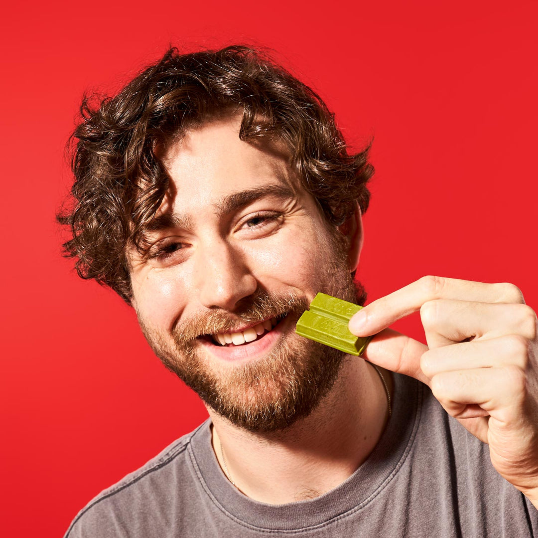 A smiling man with curly hair eats a piece of matcha Nestle Japan Rich Green Tea Free Gift against a red background.