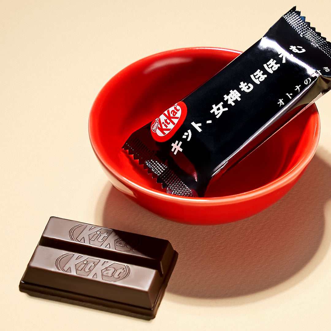 A red bowl with a wrapped Nestle Japan Kit Kat bar labeled in Japanese, beside an unwrapped Nestle Japan Kit Kat: Dark Chocolate on a light beige surface in recyclable packaging.