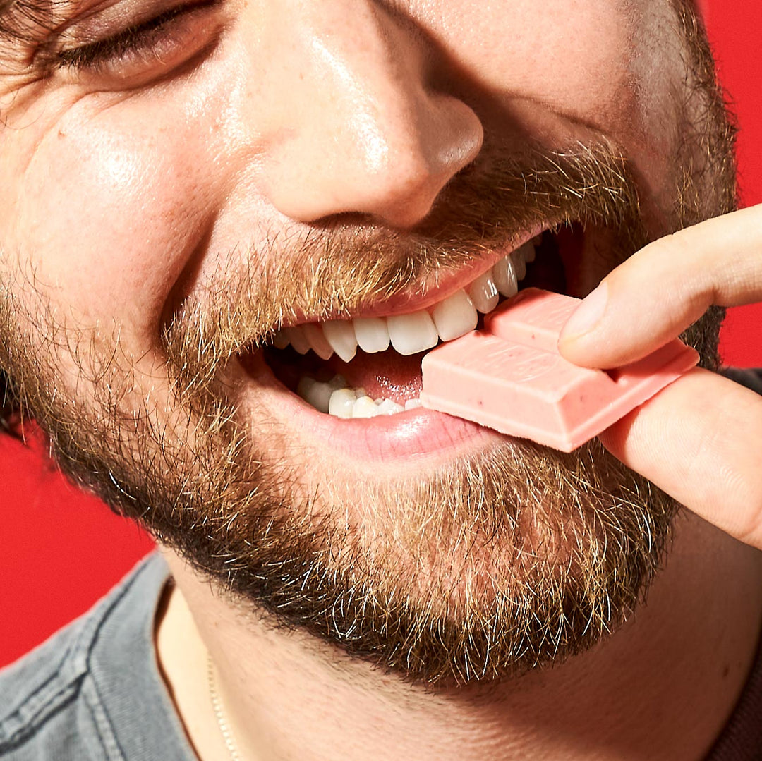 A close-up of a smiling man with a beard eating a piece of bittersweet Nestle Japan Japanese Kit Kat: Strawberry Otona no Amasa against a red background.
