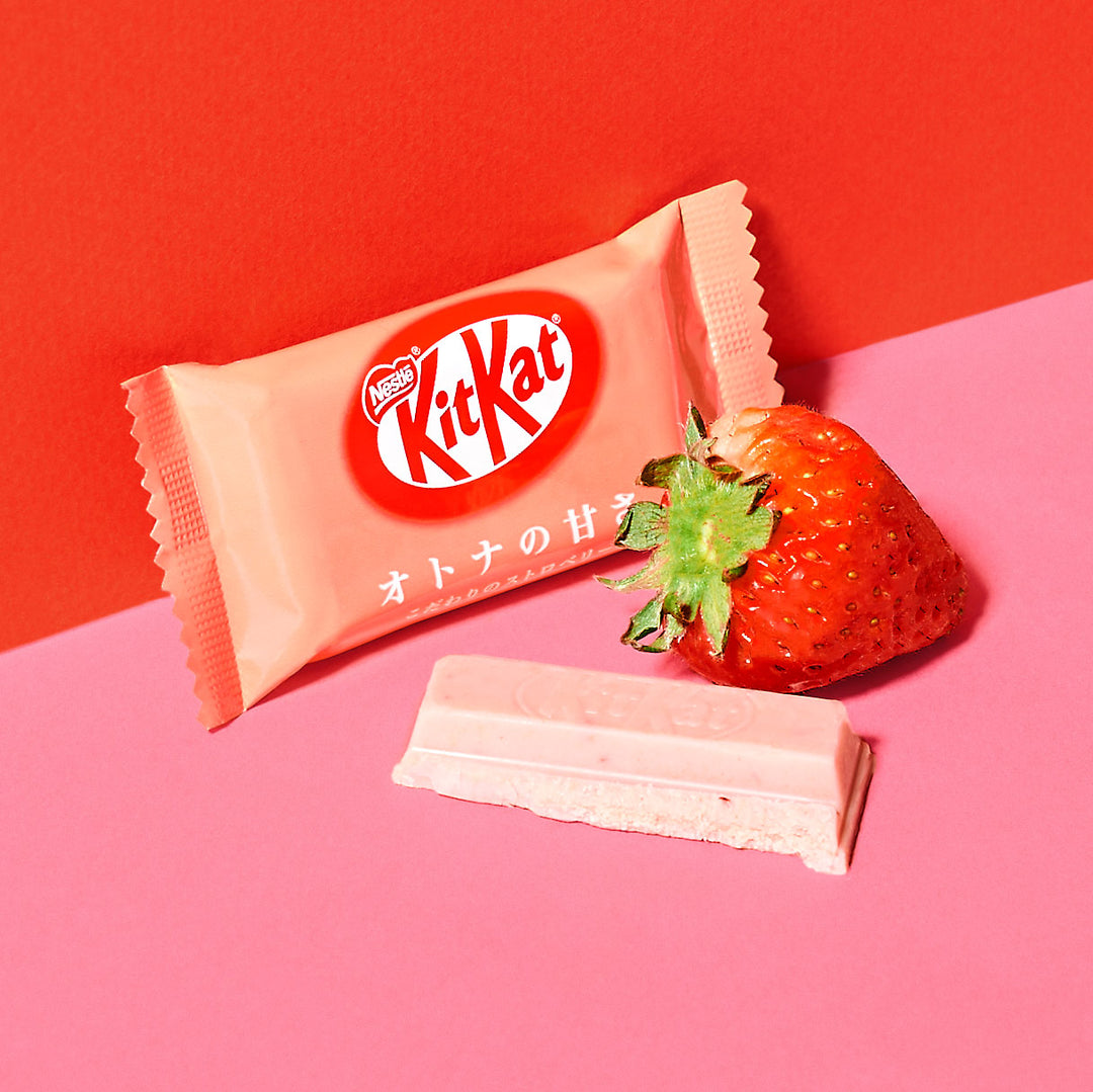 A bittersweet strawberry-flavored Nestle Japan Kit Kat: Strawberry Otona no Amasa bar next to its packaging and a whole strawberry on a vibrant pink and red background.