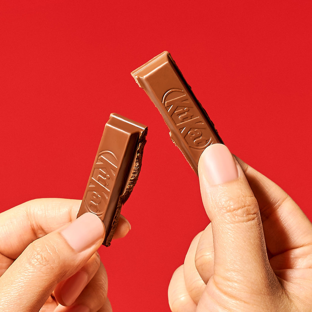 Two hands holding broken pieces of a Nestle Japan Japanese Kit Kat: Original Chocolate bar against a red background.
