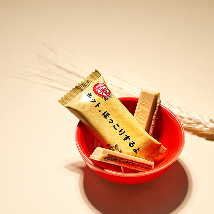 A Nestle Japan Japanese Kit Kat: Whole Wheat Cookie in a red bowl with a golden wrapper, displayed next to dried wheat stalks on a beige surface, appealing to health-conscious snackers.