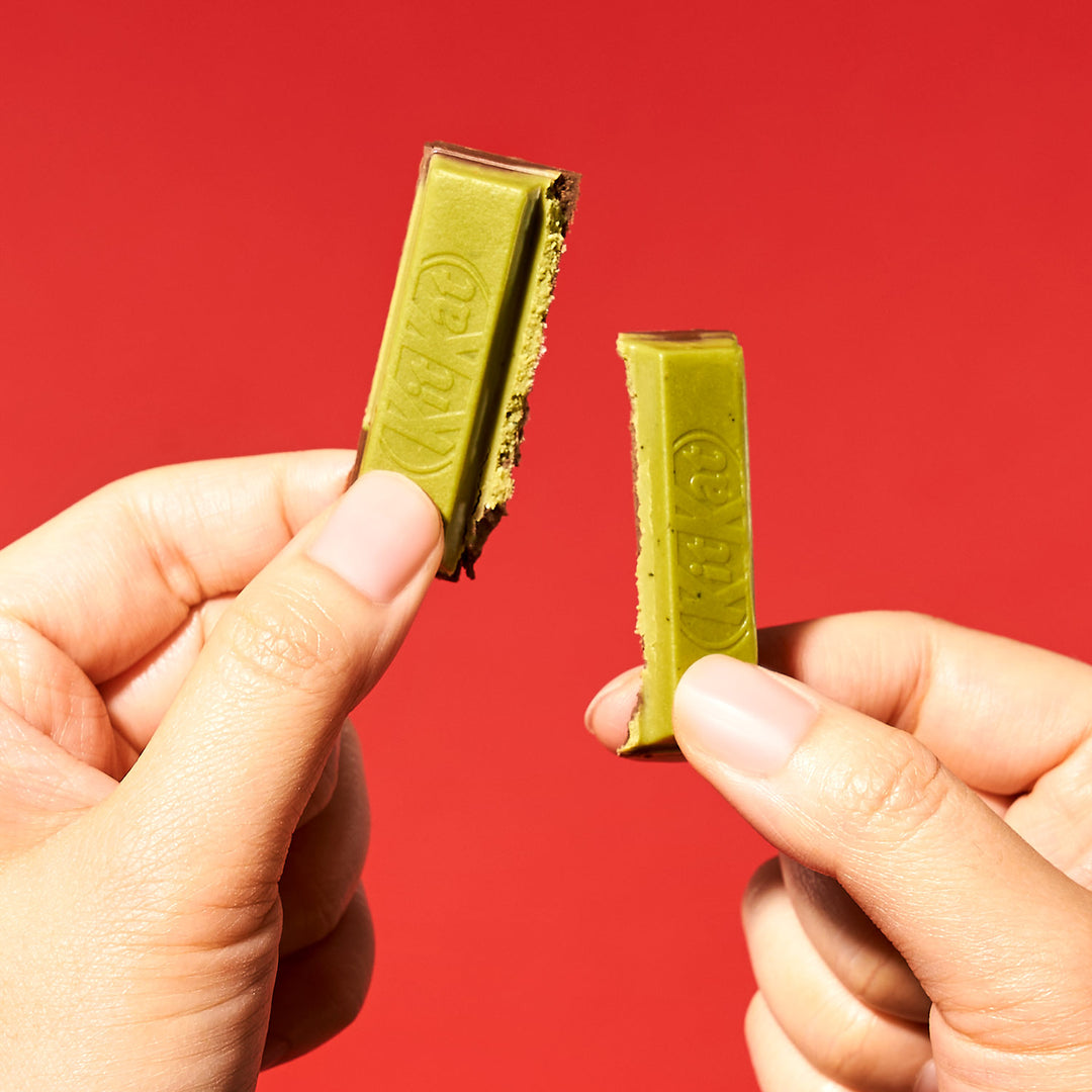Two hands holding a snapped Green Tea Double Layer Japanese Kit Kat bar from Nestle Japan against a red background.