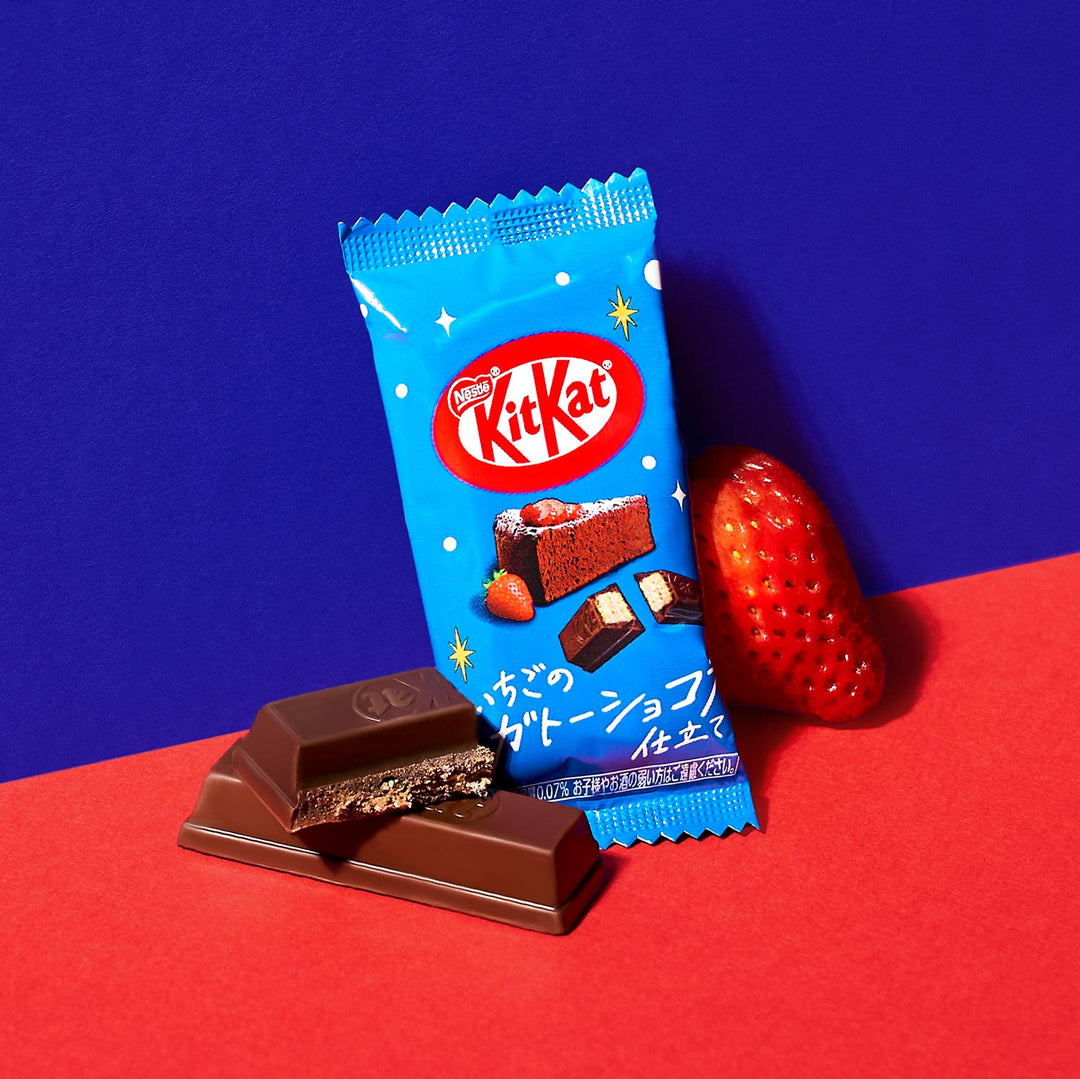 A package of Nestle Japan's Japanese Kit Kat: Strawberry Chocolate Cake with a partially unwrapped chocolate bar and a fresh strawberry on a blue and red background.