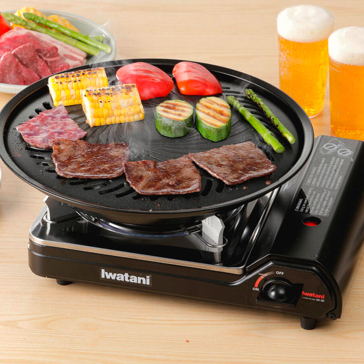 Iwatani Portable Butane Stove featuring a magnetic locking system, with various foods cooking, including steak, corn, tomatoes, and asparagus, accompanied by two glasses of beer.