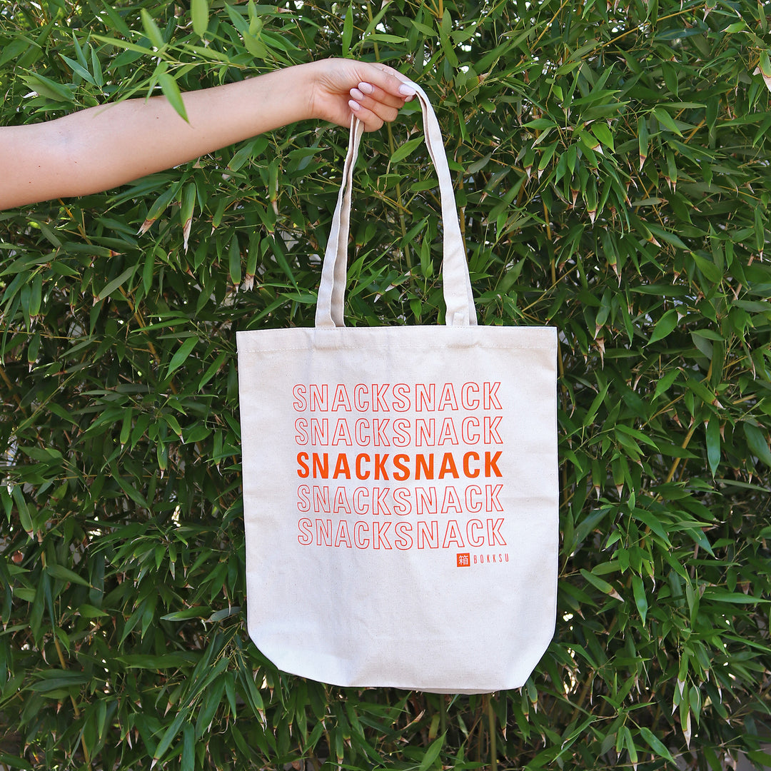 A woman holding a Bokksu Snack Tote Bag that says snacksack.